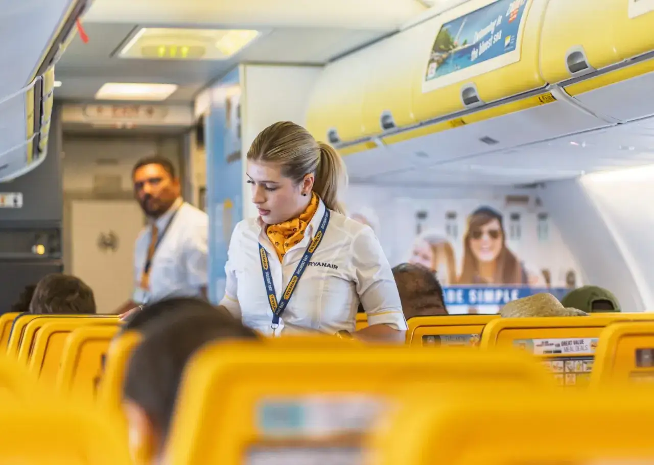 Uśmiechnięta stewardessa Ryanair w żółtym szaliku i białej koszuli, gotowa do obsługi pasażerów. Czy Ryanair jest bezpieczny? Personel dba o komfort podróży.