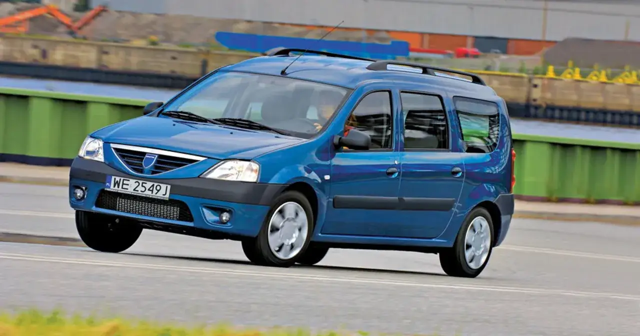 Niebieski Dacia Logan MCV II jedzie po drodze, z widokiem na nabrzeże i budynki w tle.