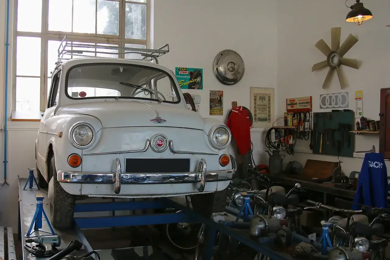 Biały Fiat 600 na podnośniku w warsztacie, gotowy do napraw. W tle narzędzia i części, a nad nimi oświetlenie kanału samochodowego.