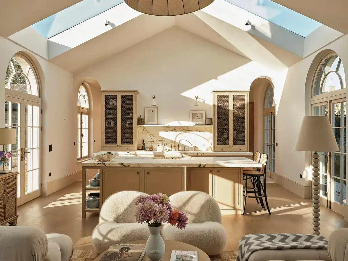 Sunlight streams through a large skylight into a bright, airy modern farmhouse kitchen. Arched windows and doors, a marble island, and plush seating create a welcoming atmosphere.