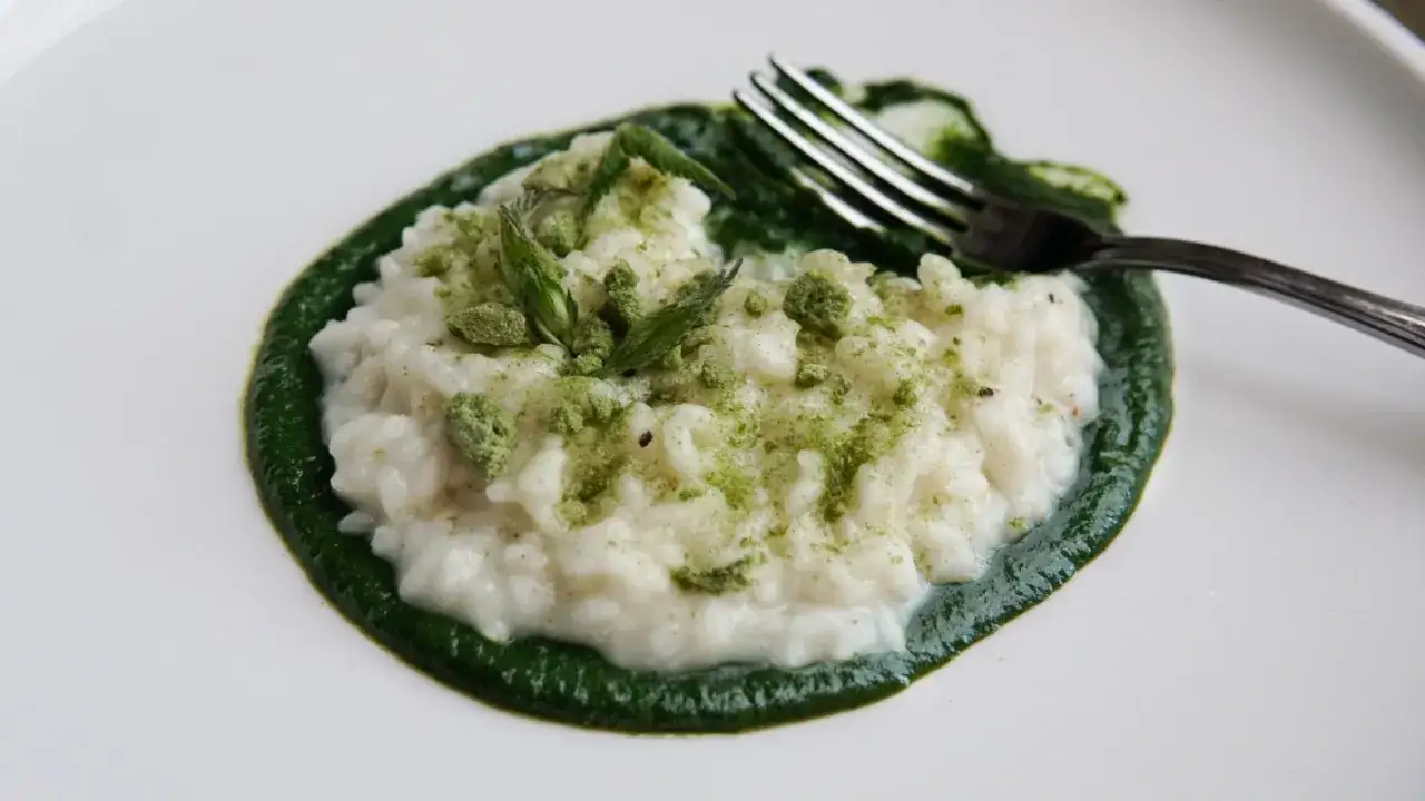Un cremoso risotto con ortiche, guarnito con polvere verde e germogli freschi. Un piatto primaverile che celebra i sapori della natura.