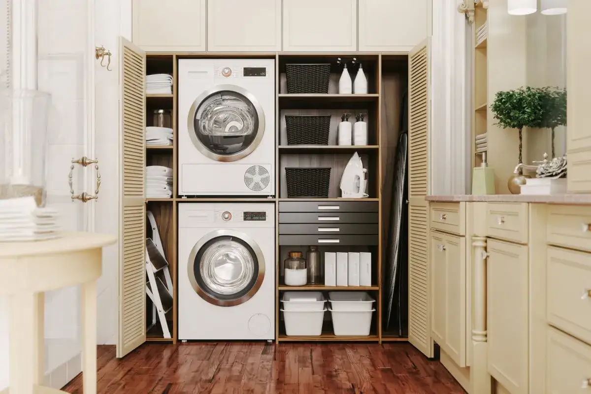 Un lave-linge et un sèche-linge superposés dans une buanderie bien rangée, avec des paniers, des bouteilles et un fer à repasser sur des étagères.