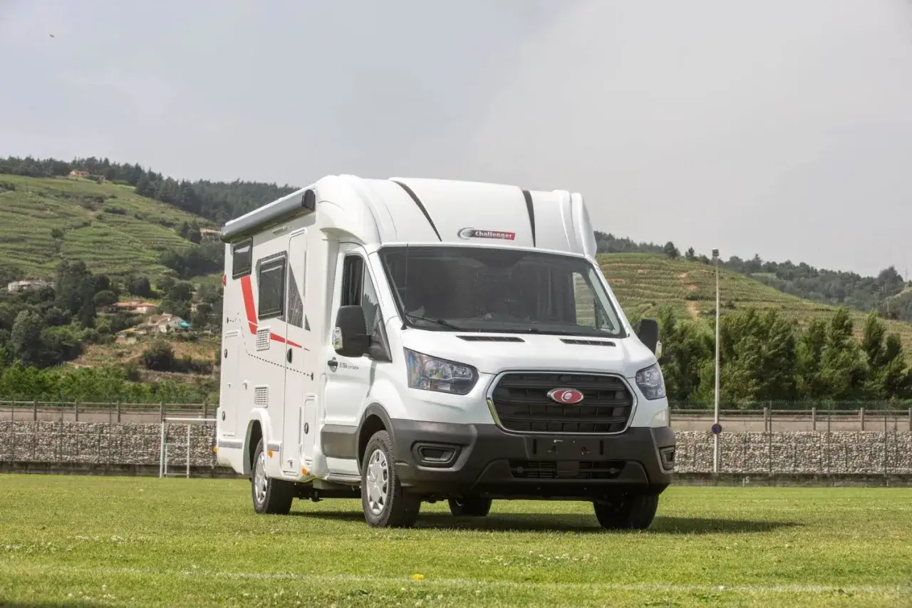 Porteur Ford camping-car blanc, prêt pour l'aventure sur un terrain de sport verdoyant, avec des collines en arrière-plan.