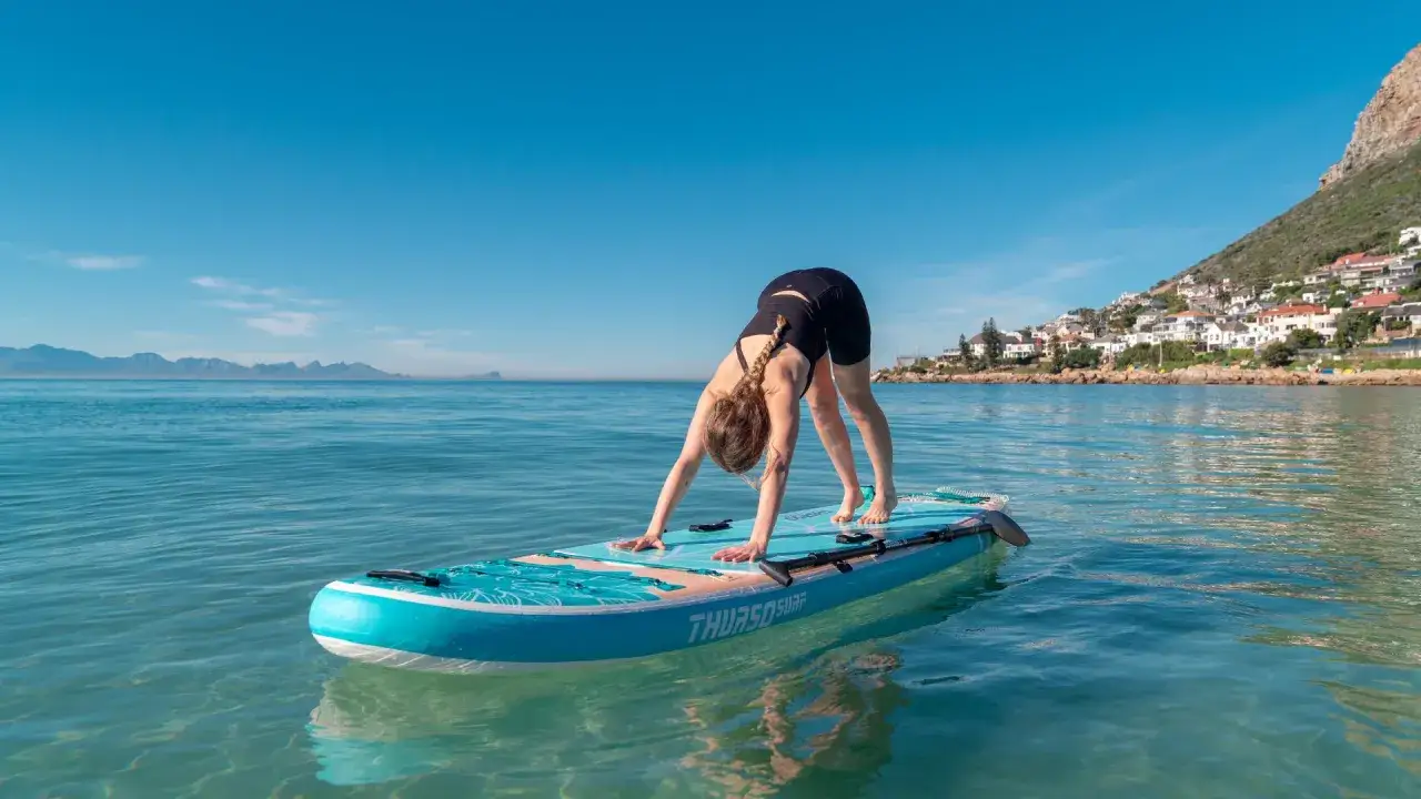 Kvinna g&ouml;r yoga p&aring; SUP-br&auml;da i lugnt vatten. Ned&aring;tg&aring;ende hund-position med berg och hus i bakgrunden.