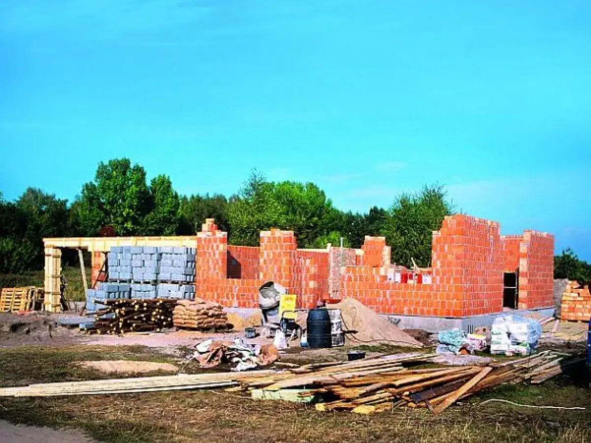 Budowa domu systemem gospodarczym w toku. Wzniesione mury z cegły, sterty materiał&oacute;w budowlanych: pustaki, drewno, worki z cementem.