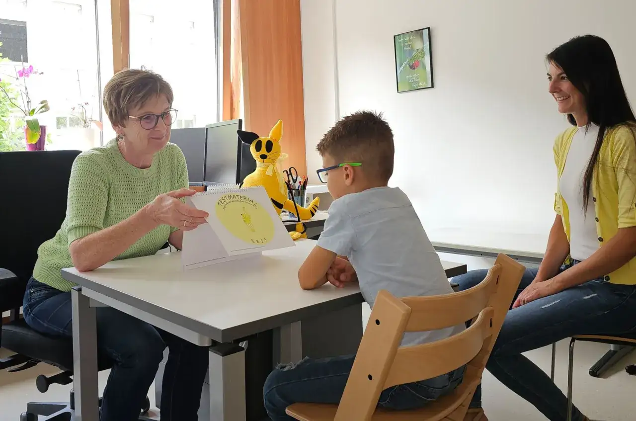 Frau zeigt Kind und Mutter ein Heft mit dem Titel
