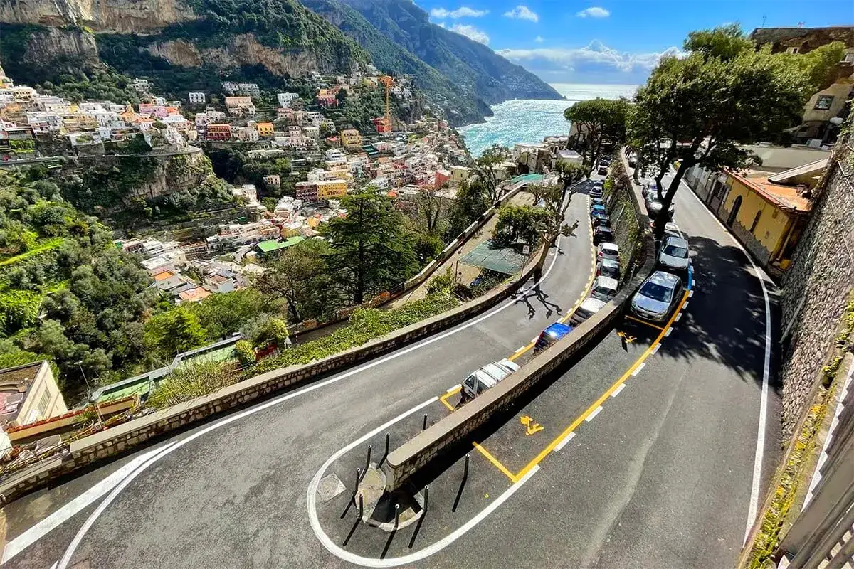 A winding road with parked cars overlooks the colorful houses of Positano, a scenic route to consider when planning how to get from Amalfi to Naples.