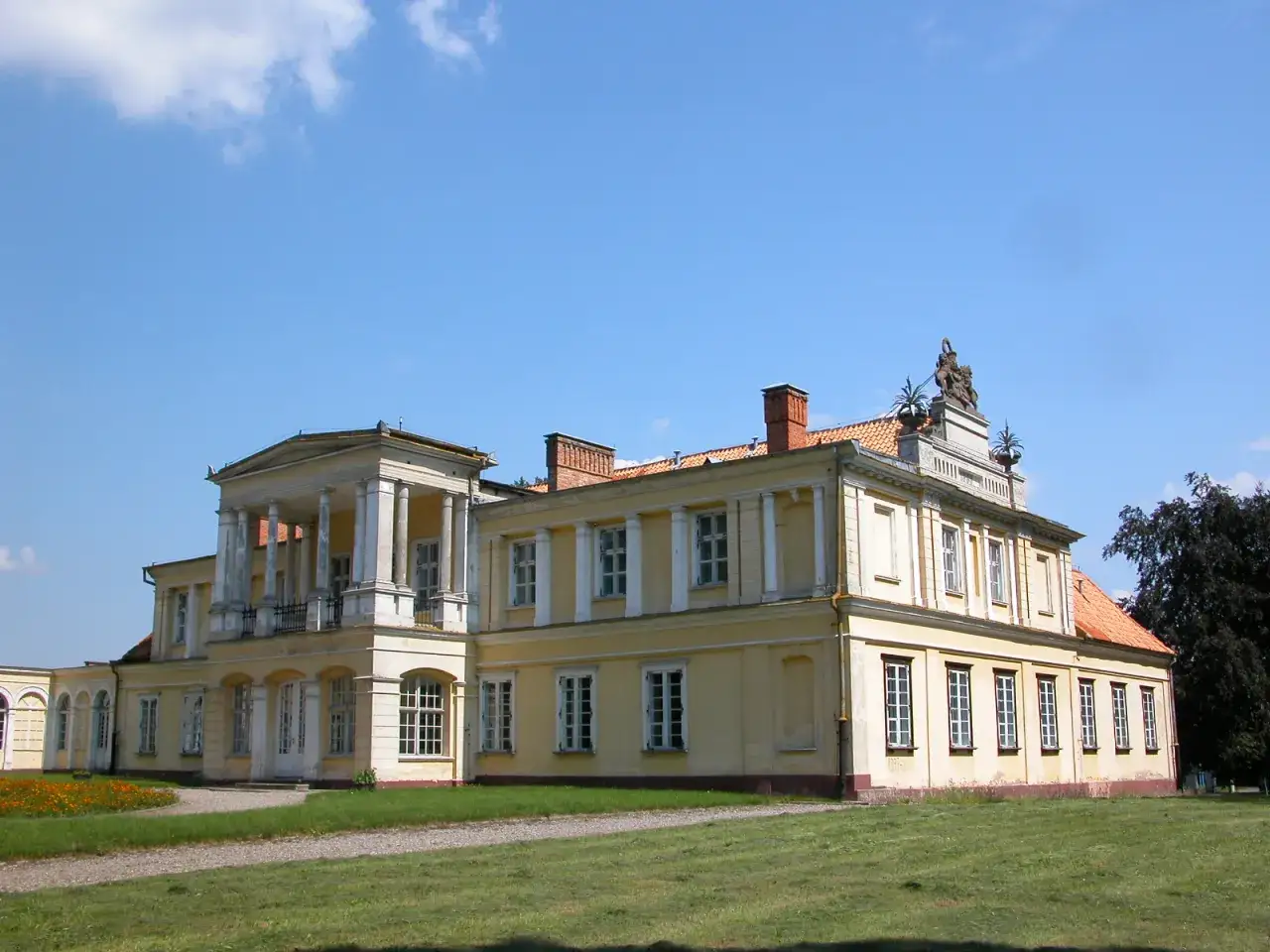 Pałac w Waplewie, żółty, z kolumnami i balkonem, otoczony zielonym trawnikiem i kwitnącymi kwiatami.