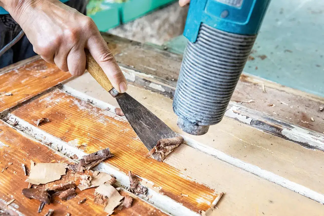 Mit einem Heißluftföhn und einem Spachtel wird alter Lack von Holz entfernt.