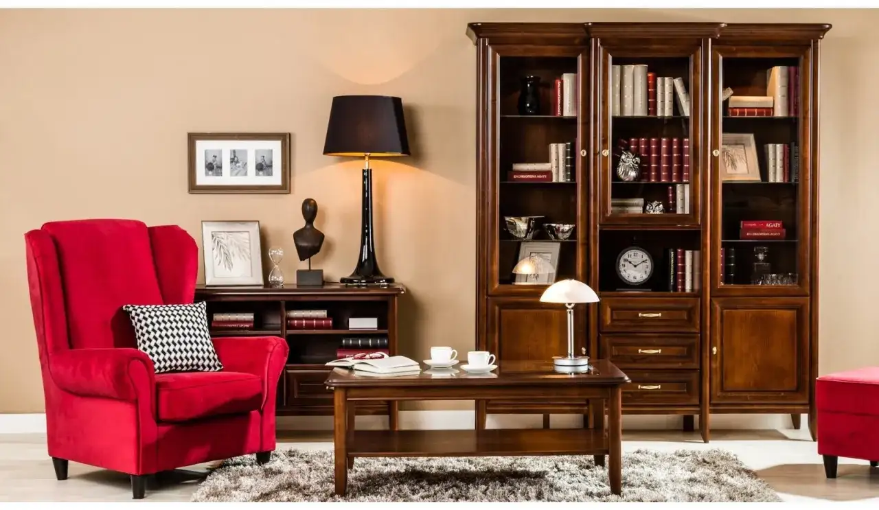 Elegancki salon w stylu tradycyjnym. Czerwony fotel, drewniane meble, regały pełne książek i stylowe lampy tworzą przytulną atmosferę.