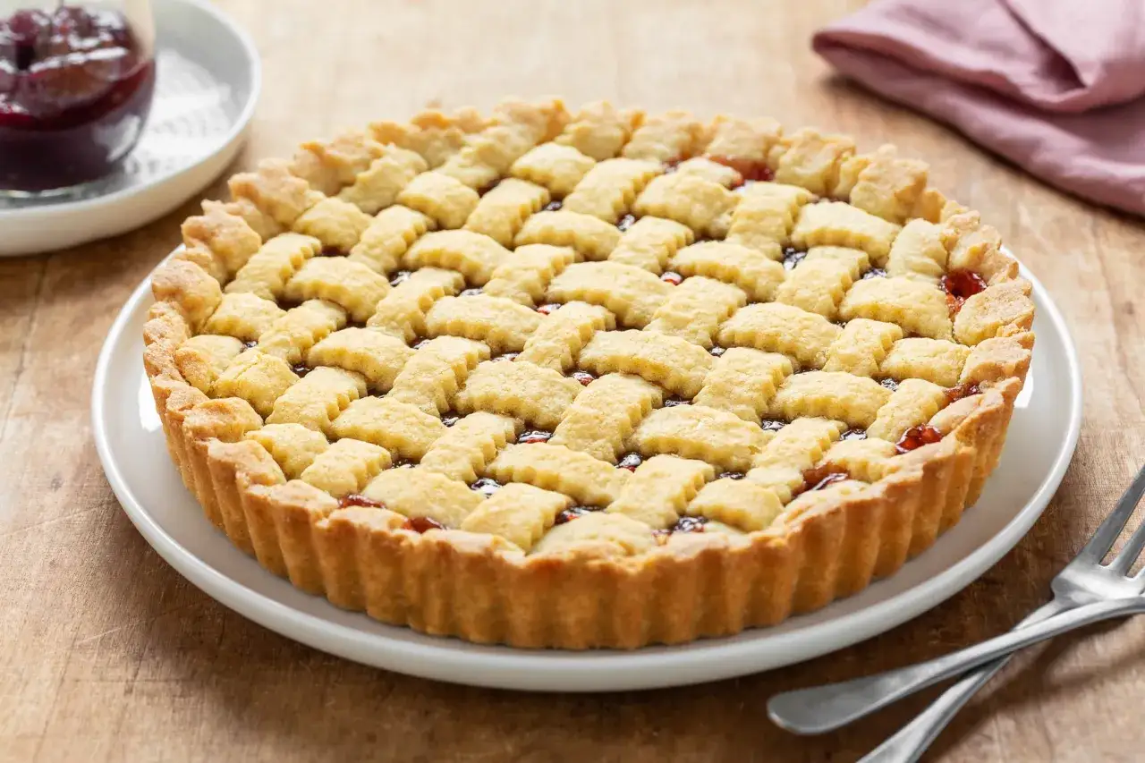 Crostata ricetta tradizionale con marmellata di frutti rossi, decorata con una griglia di pasta frolla dorata.