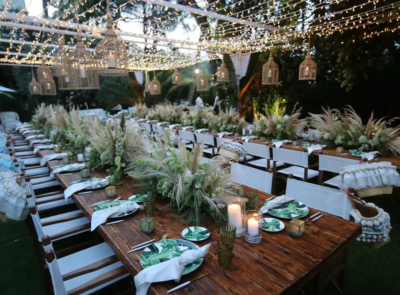 Tavola lunga apparecchiata per un matrimonio in campagna, con centrotavola di fiori e pampas, luci soffuse e cesti decorativi.