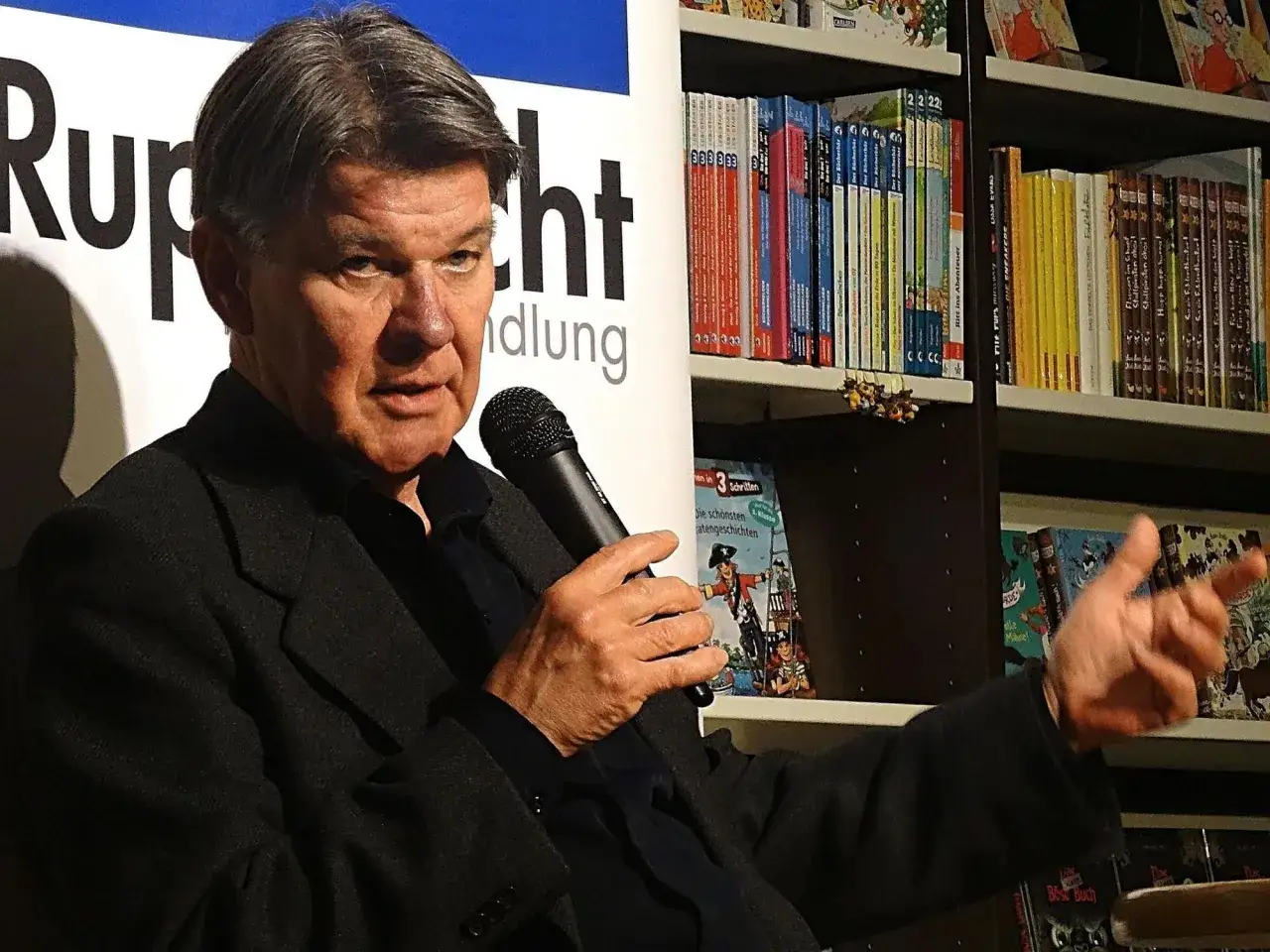 Udo Lielischkies spricht in ein Mikrofon, im Hintergrund Bücherregale.