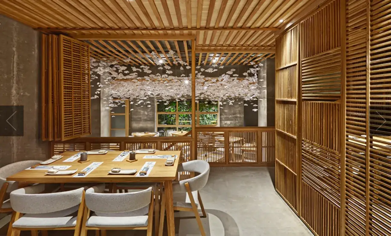 Interior de restaurante japonés con mesas de madera, sillas grises y una decoración de techo con flores blancas. La barra de sushi está lista para recibir a los comensales.