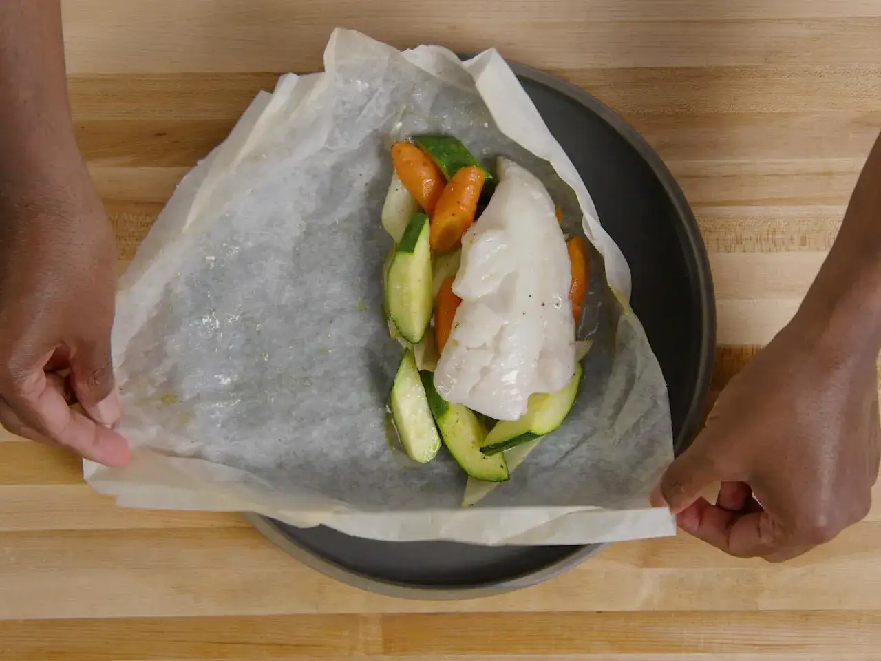Préparation d'un poisson en papillote avec des légumes frais. Les mains replient délicatement le papier sulfurisé.