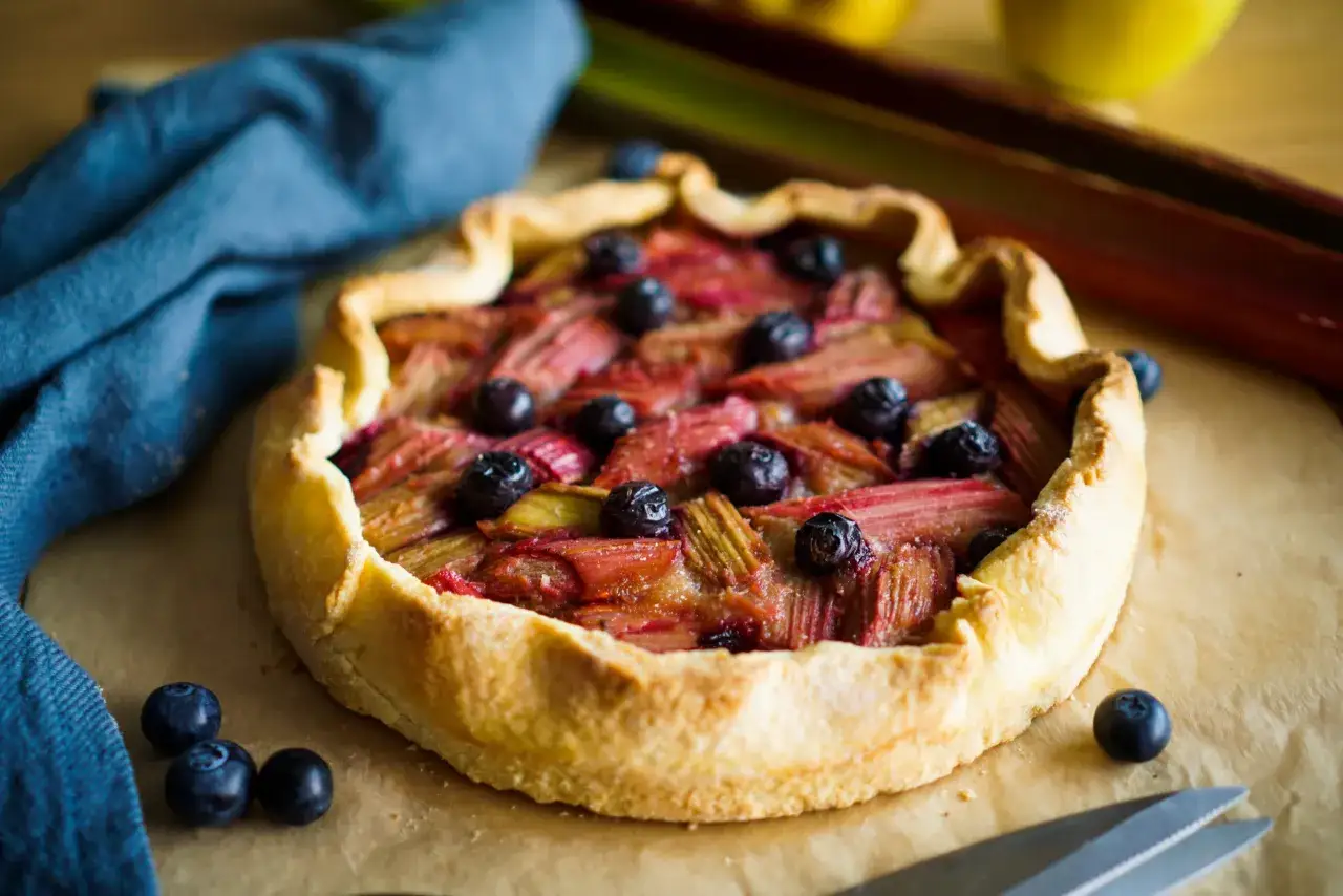 Tarte rustique aux fruits rouges, rhubarbe et pommes, garnie de myrtilles fraîches. Un délice gourmand.