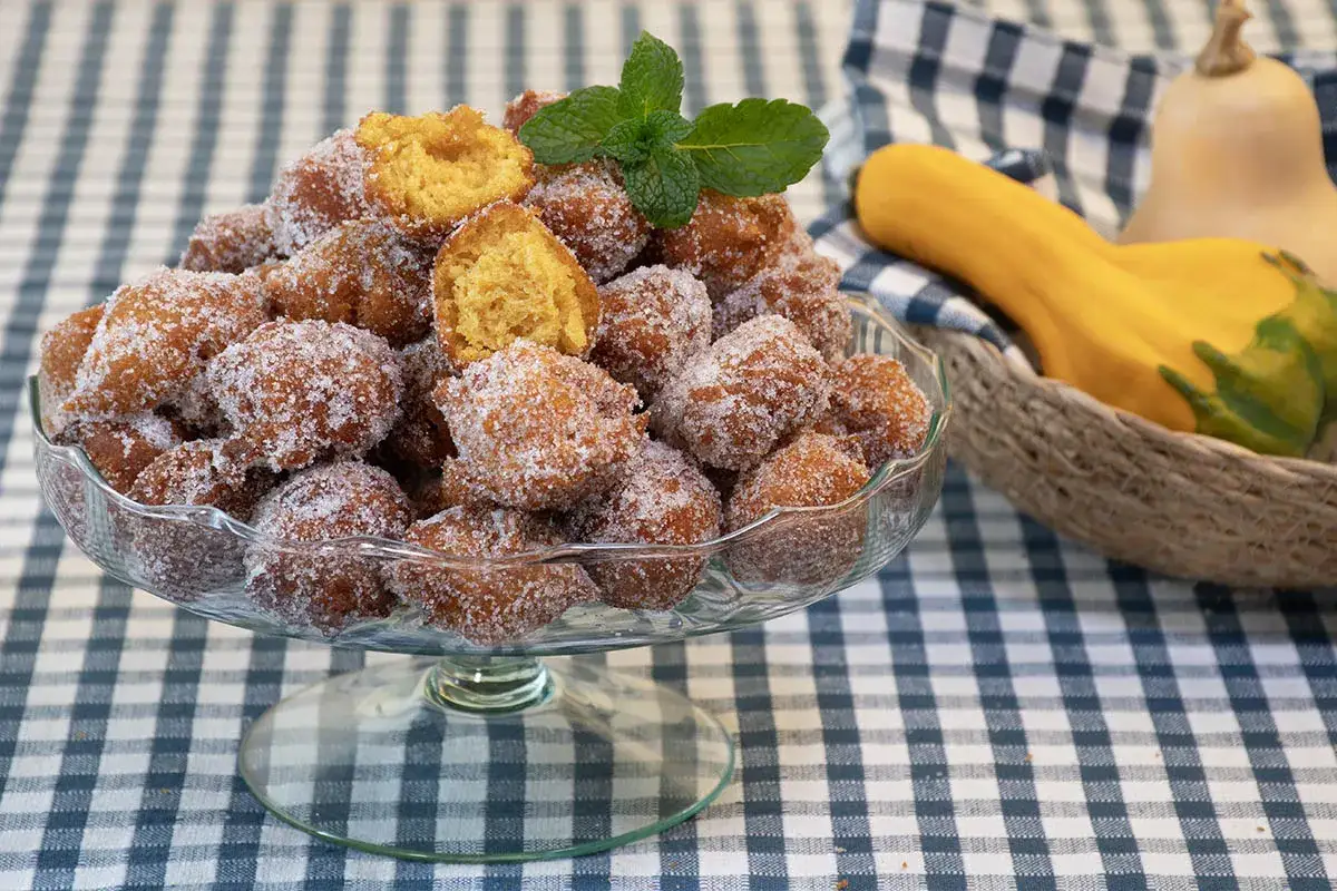 Un taz&oacute;n lleno de bu&ntilde;uelos de calabaza cubiertos de az&uacute;car, adornados con una ramita de menta. Al fondo, calabazas en una cesta.