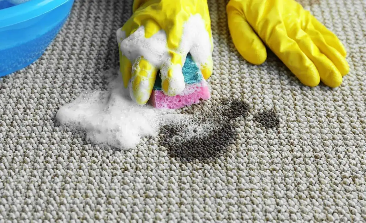 Gelbe Handschuhe schrubben mit einem Schwamm und Schaum einen Fleck auf einem hellen Teppich.