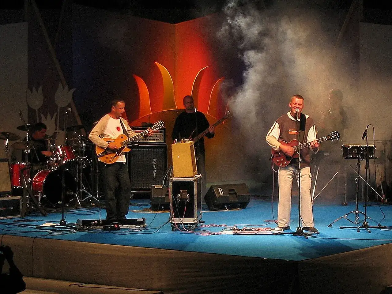 Tercet gitarzystów na scenie, jeden śpiewa, w tle dym i światła.