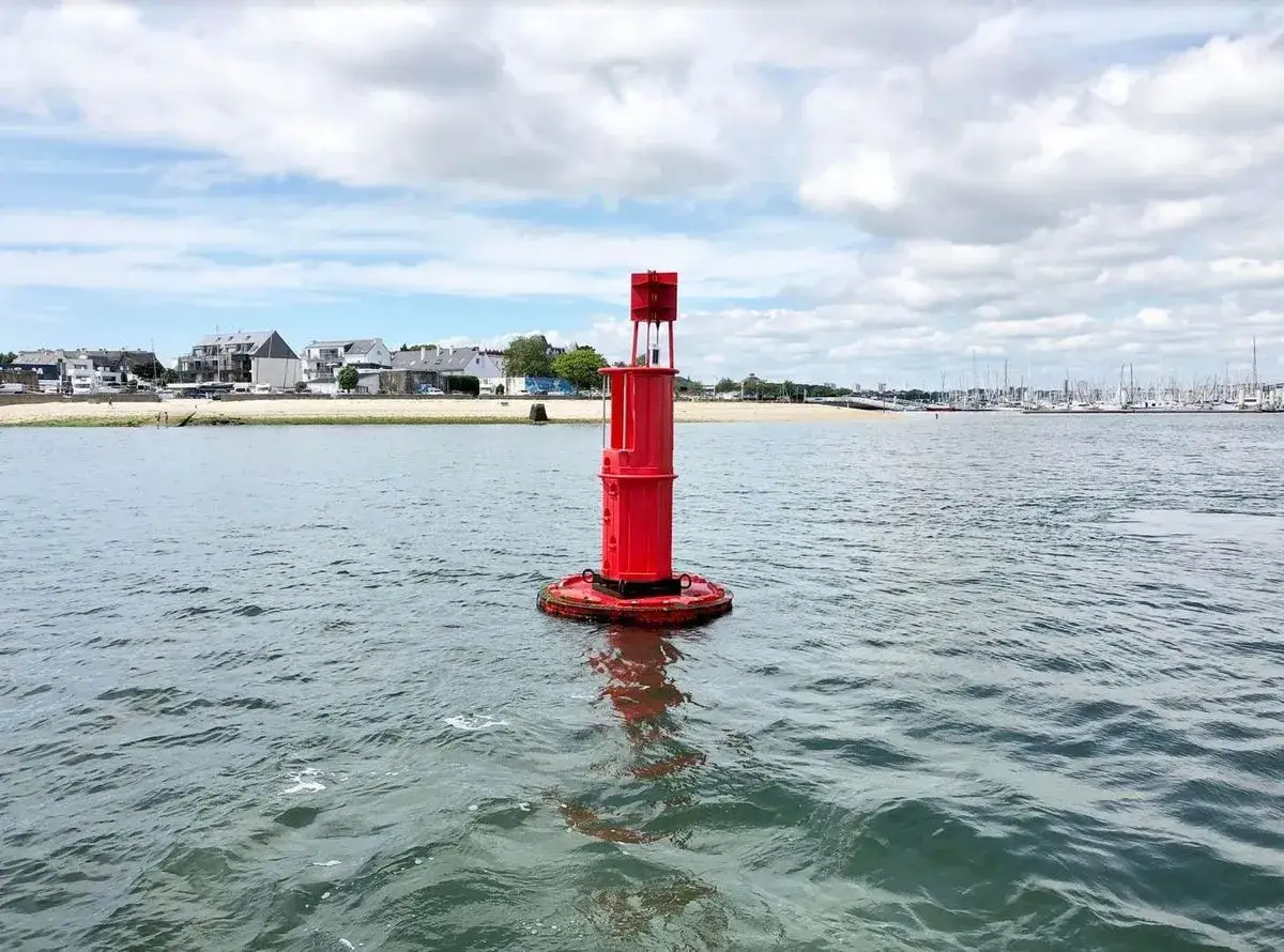 Une balise rouge imposante flotte sur l'eau, guidant les navires vers le port. Au loin, des maisons et des voiliers bordent la c&ocirc;te.