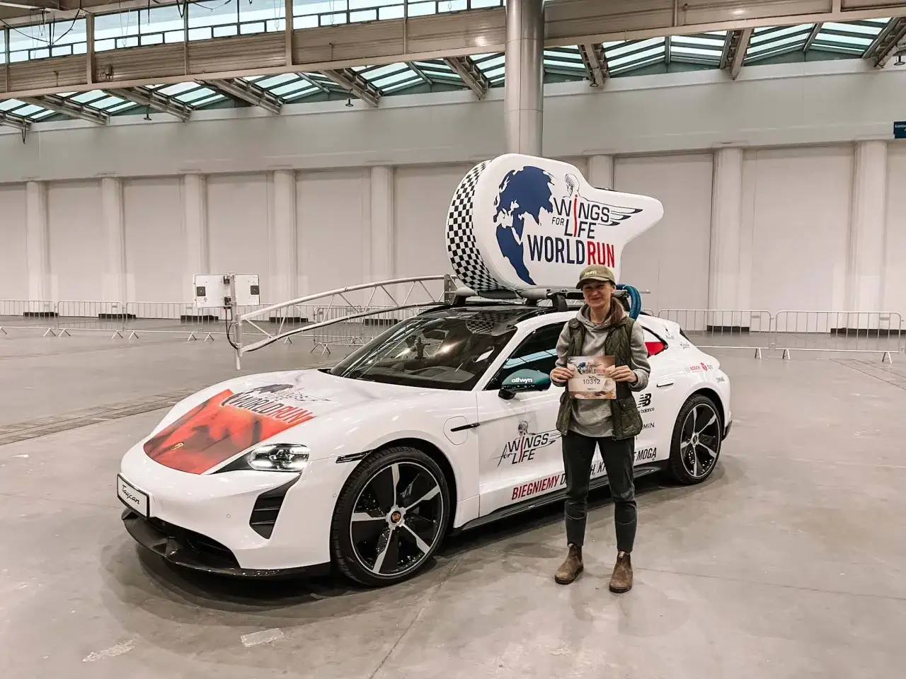 Kobieta z numerem startowym przed Porsche z logo Wings for Life World Run. Gotowa na bieg w 2026!