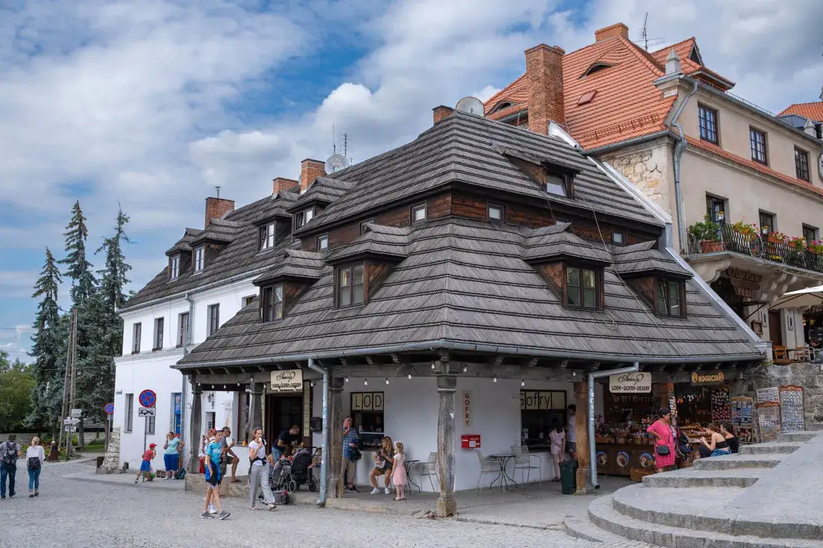 Kazimierz Dolny rynek: urokliwe kamienice z drewnianymi dachami, ludzie spacerują, kupują lody i pamiątki.
