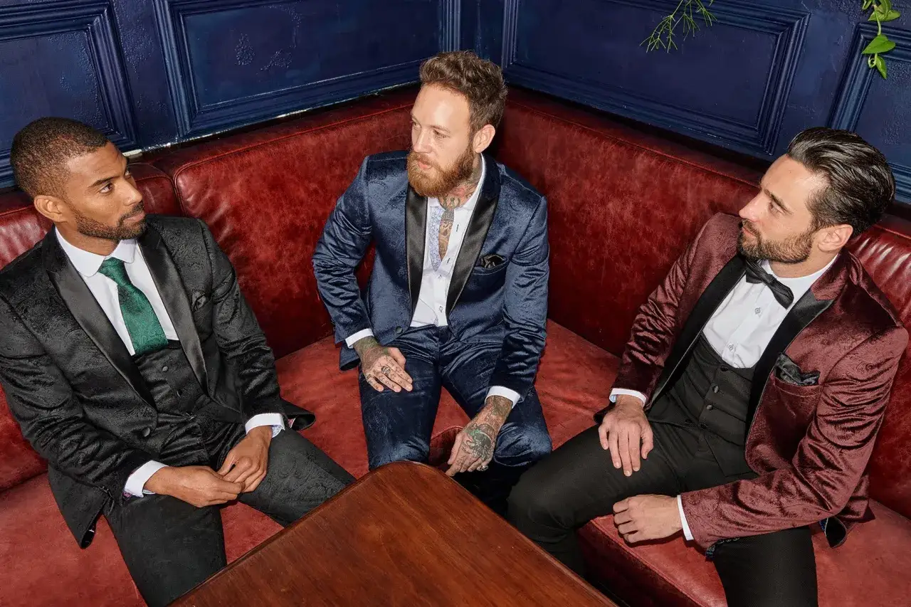 Three men in stylish, dressy attire for men: a black patterned suit with a green tie, a blue patterned suit, and a burgundy patterned suit with a bow tie, all seated on a red leather couch.