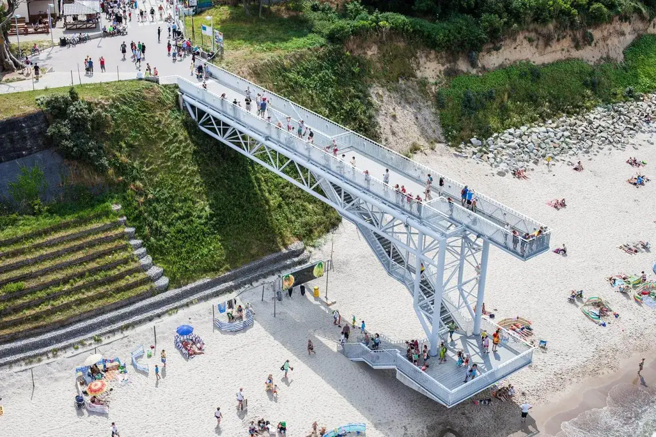 Nowa kładka w Rewalu to jedna z głównych atrakcji, łącząca plażę z promenadą. Widać tu spacerujących ludzi i plażowiczów.