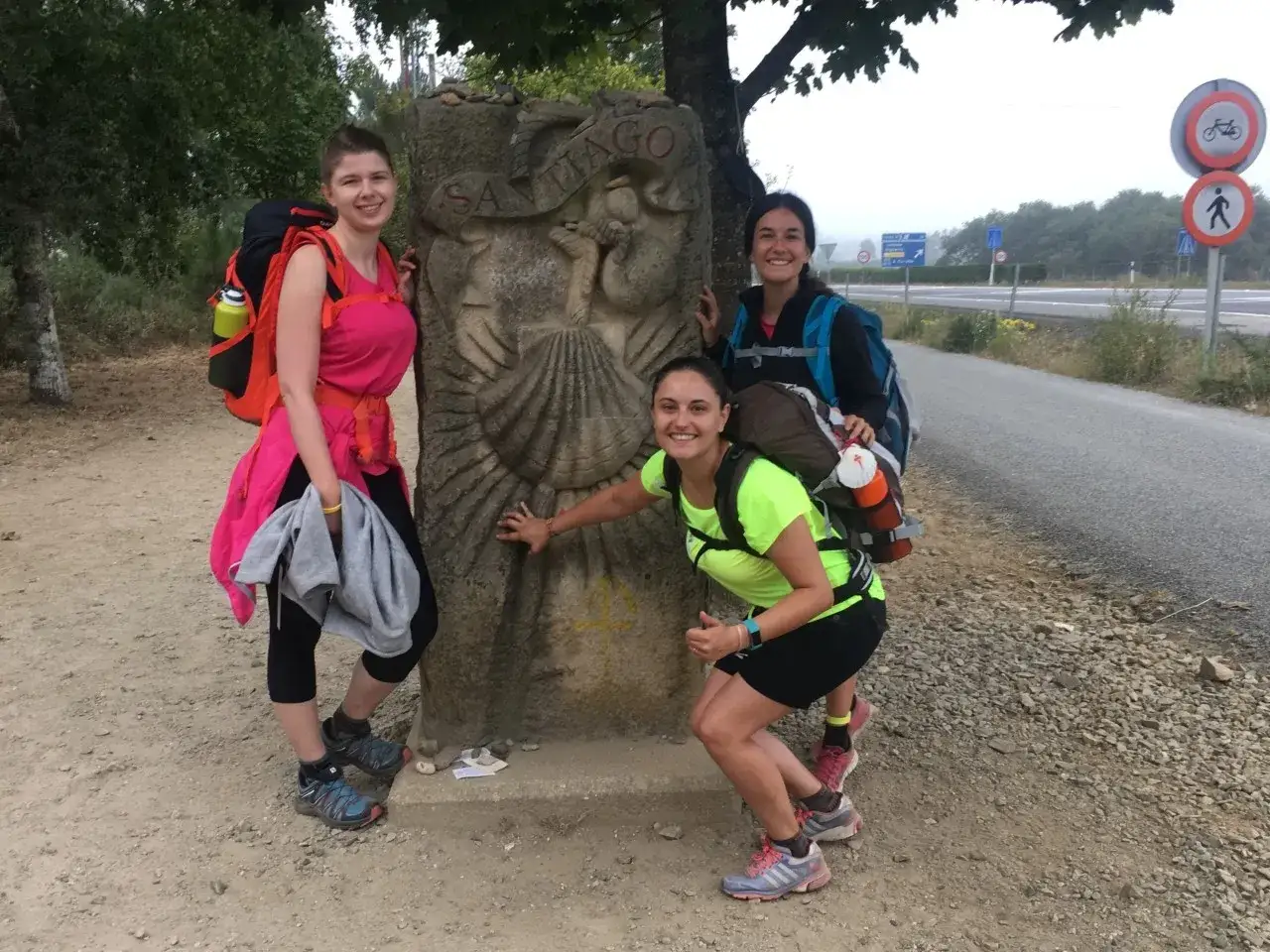 Tres peregrinas sonr&iacute;en junto a un moj&oacute;n del Camino de Santiago. El tama&ntilde;o de su mochila es perfecto para el camino.