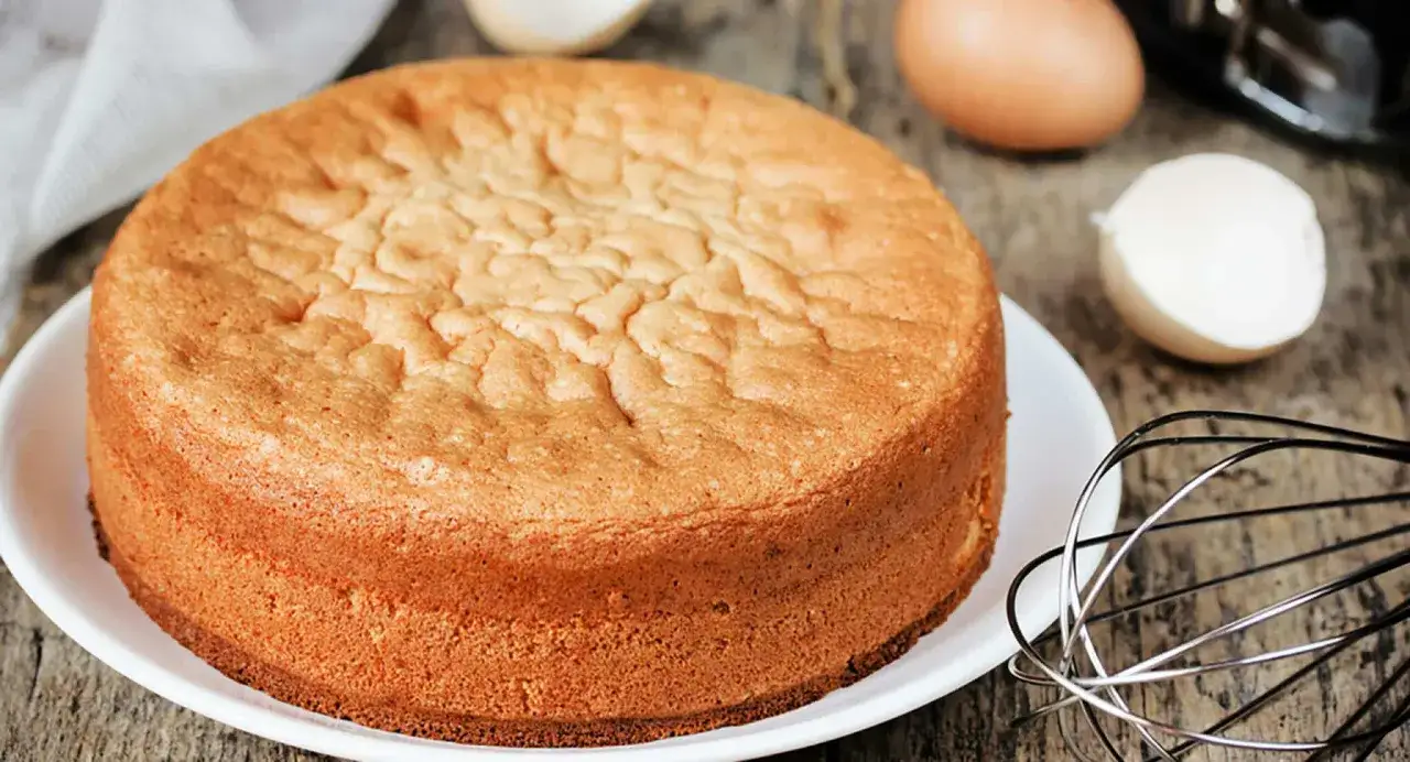 Ein goldbrauner Biskuitkuchen auf einem Teller, daneben Eier und ein Schneebesen. Perfekte Biskuit Backzeit und Temperatur sind entscheidend f&uuml;r dieses Ergebnis.