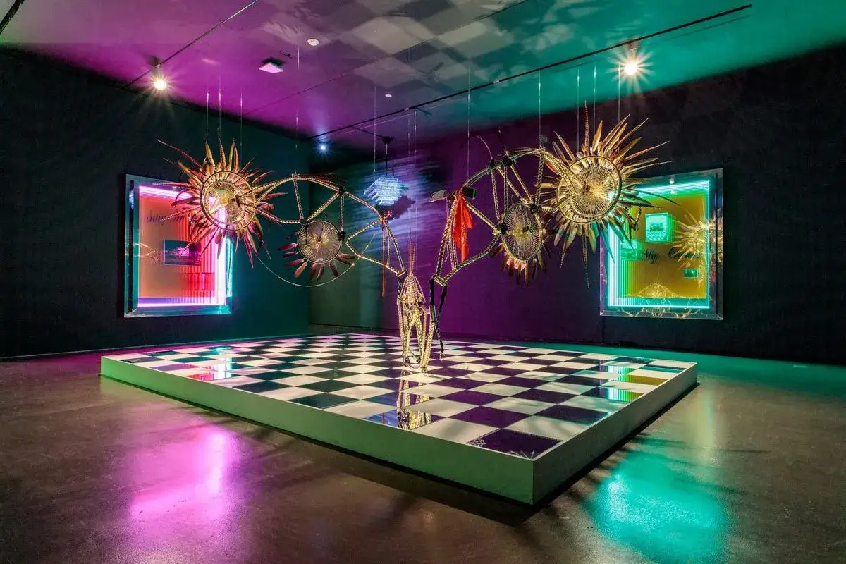 A fantastical, golden, winged sculpture hangs above a checkered floor in this installation made in LA Hammer.