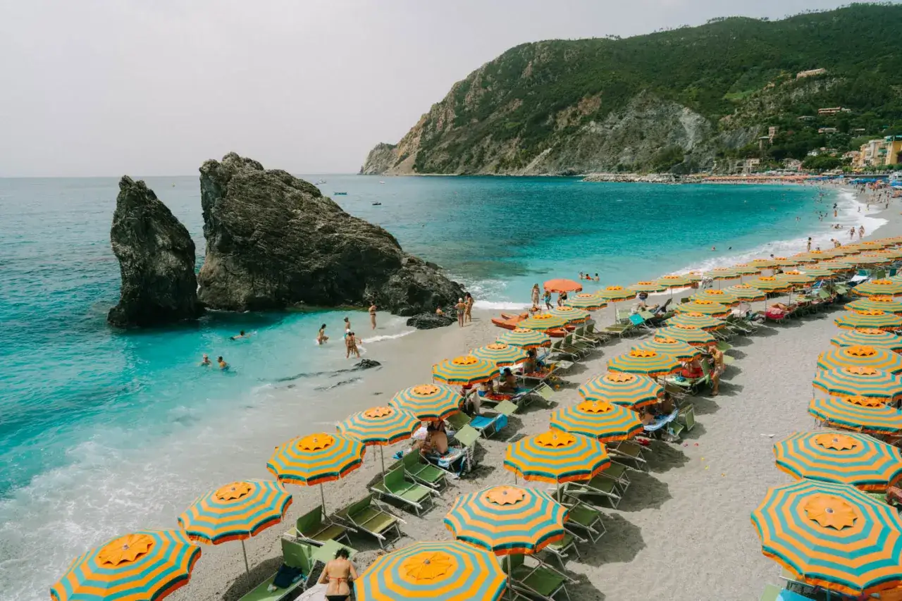 Plage animée de la **riviera italienne où aller** pour se baigner et se détendre sous des parasols colorés, avec des rochers imposants et une mer turquoise.