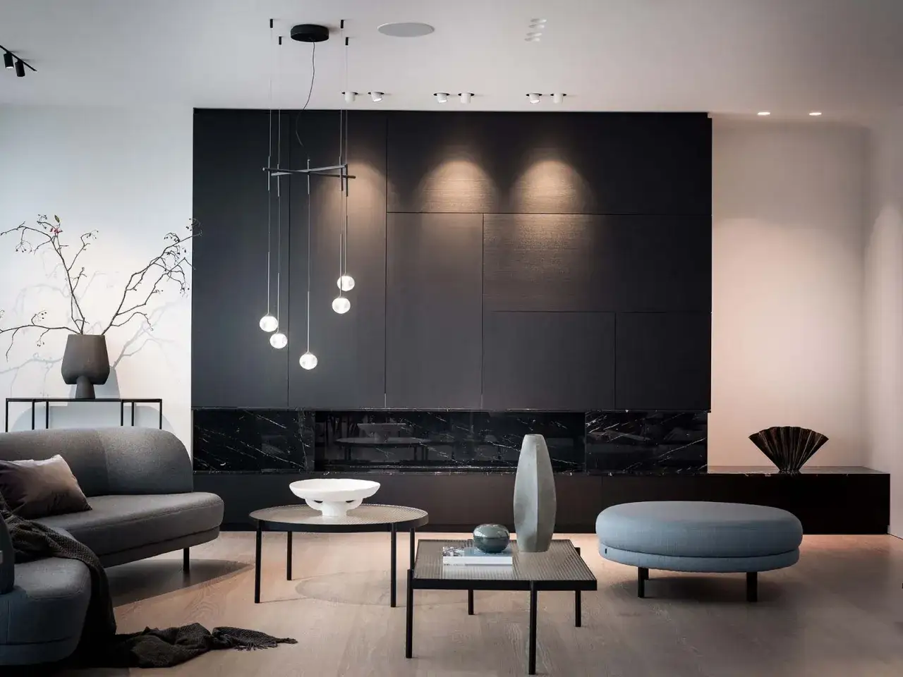 Ett lyxigt vardagsrum med modern design. En grå soffa, runda soffbord och en stor blå puff. Bakom syns en svart vägg med en eldstad och en taklampa med klotformade ljuskällor.