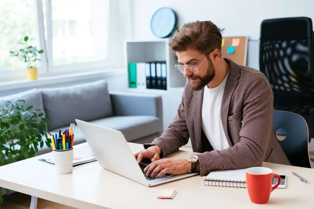 Homme concentré sur son ordinateur portable, prêt à gagner de l'argent en écrivant. Bureau moderne avec tasse rouge et stylos colorés.