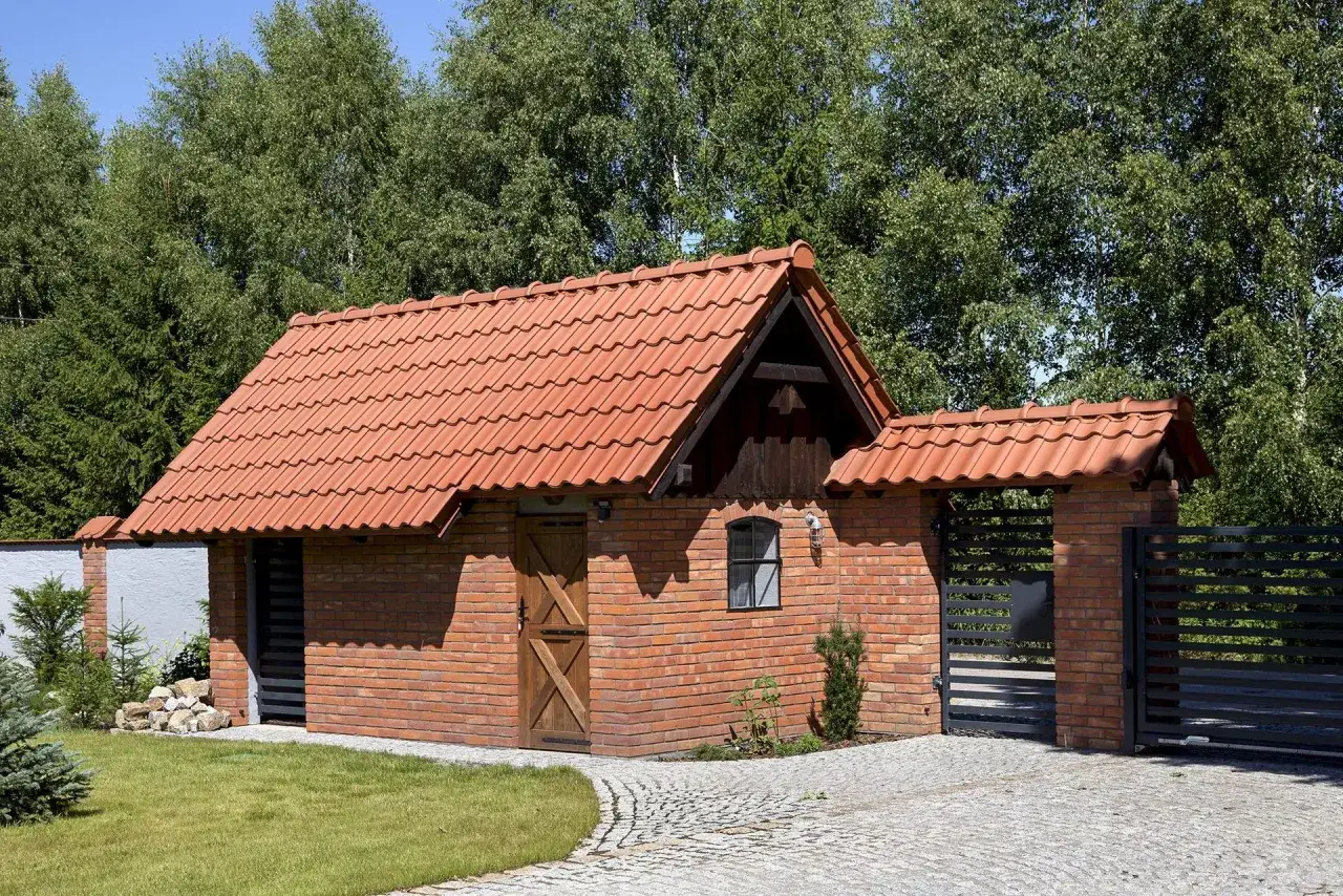 Przekształcenie budynku gospodarczego na domek letniskowy: ceglany budynek z czerwoną dachówką, drewniane drzwi, małe okienko, brama, otoczony zielenią.