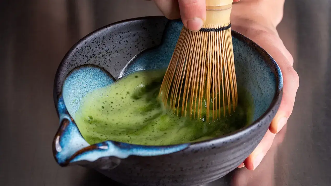 En hand vispar en grön, skummande matcha latte i en vacker keramikskål.
