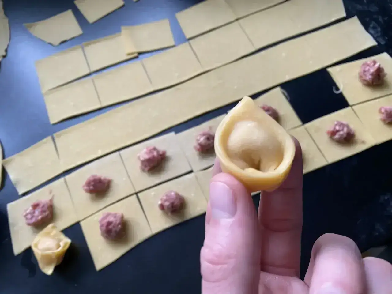 Przygotowanie tortellini in brodo: ciasto pokrojone w kwadraty, nadzienie mięsne i gotowe pierożki.