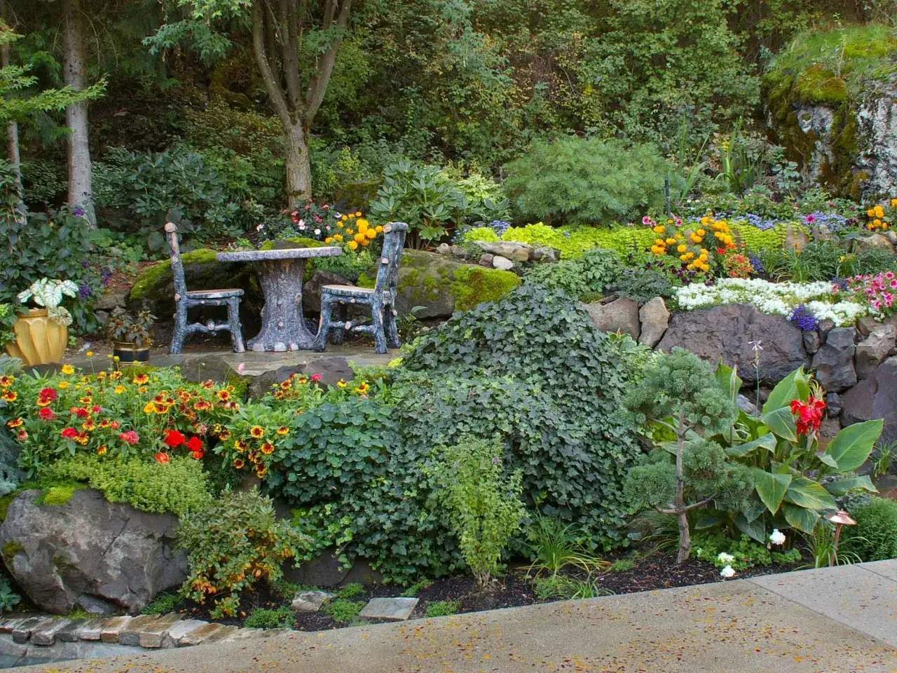 Ein idyllischer **funkien standort** mit einem Steintisch und zwei Stühlen, umgeben von üppiger Vegetation und bunten Blumen.