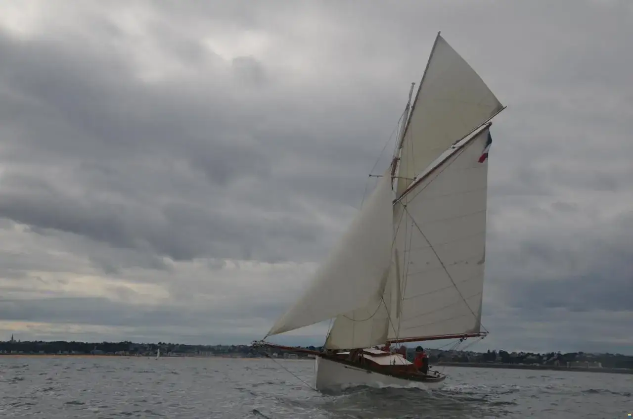 Un cotre de Carantec navigue sous un ciel menaçant, ses voiles blanches gonflées par le vent. La côte bretonne se dessine à l'horizon.
