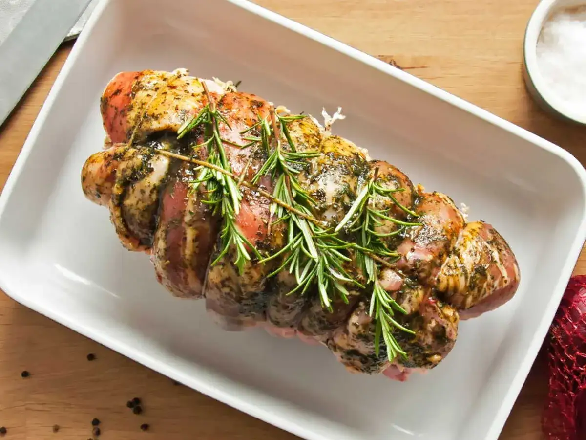 Un r&ocirc;ti de porc cru, assaisonn&eacute; et garni de romarin frais, pr&ecirc;t &agrave; &ecirc;tre cuit dans un plat blanc.