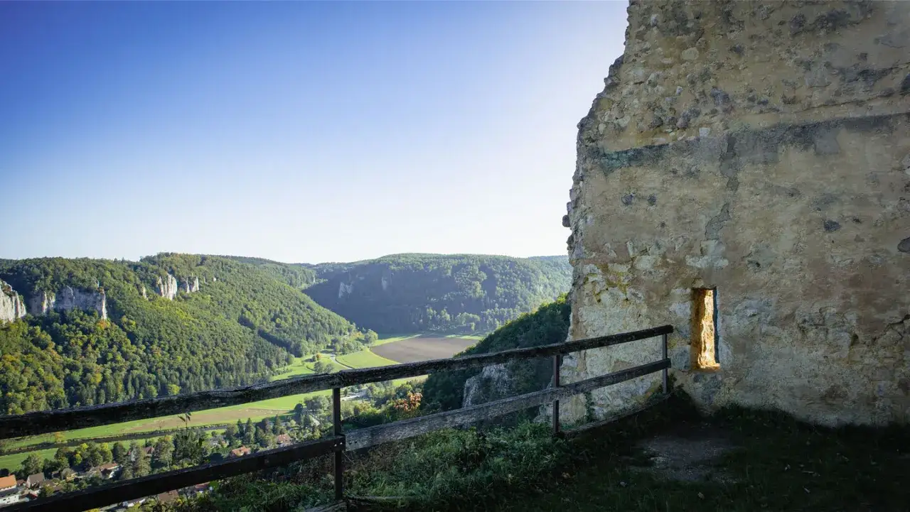 Blick von der Ruine Kallenberg über ein grünes Tal mit Felsen und Wäldern. Ein schmaler Lichtschacht erhellt die alte Mauer.
