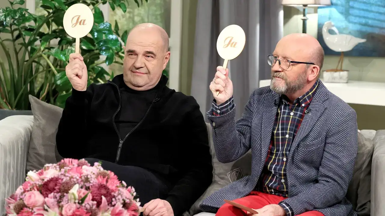 Cezary Żak i jego kolega z teatru, siedząc na kanapie, trzymają tabliczki z napisem 