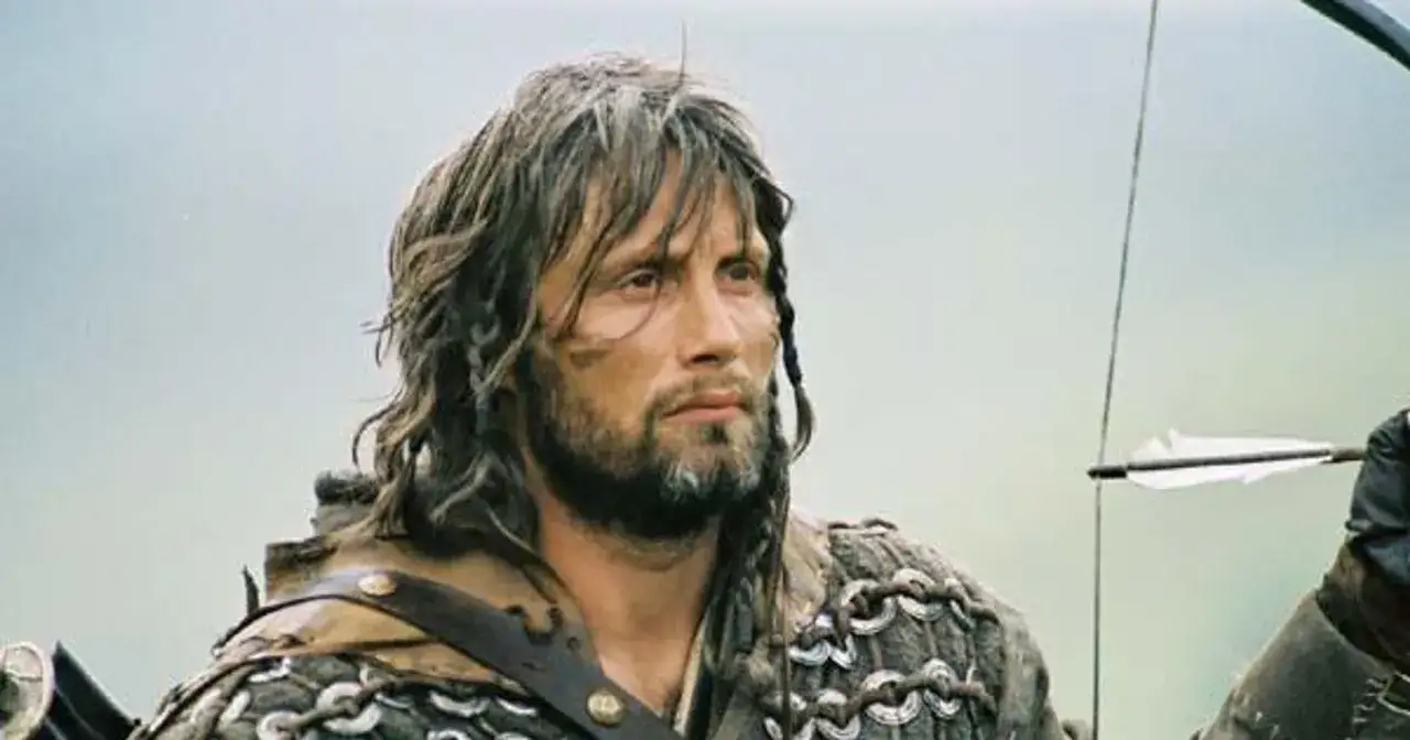 Mads Mikkelsen młody jako łucznik w zbroi, z długimi włosami i brodą, gotowy do strzału.