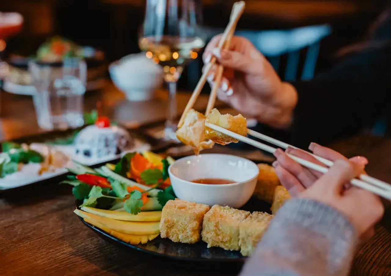 Ręce z pałeczkami zanurzają chrupiące kąski w sosie. Na talerzu qb restauracji świeże warzywa i owoce.