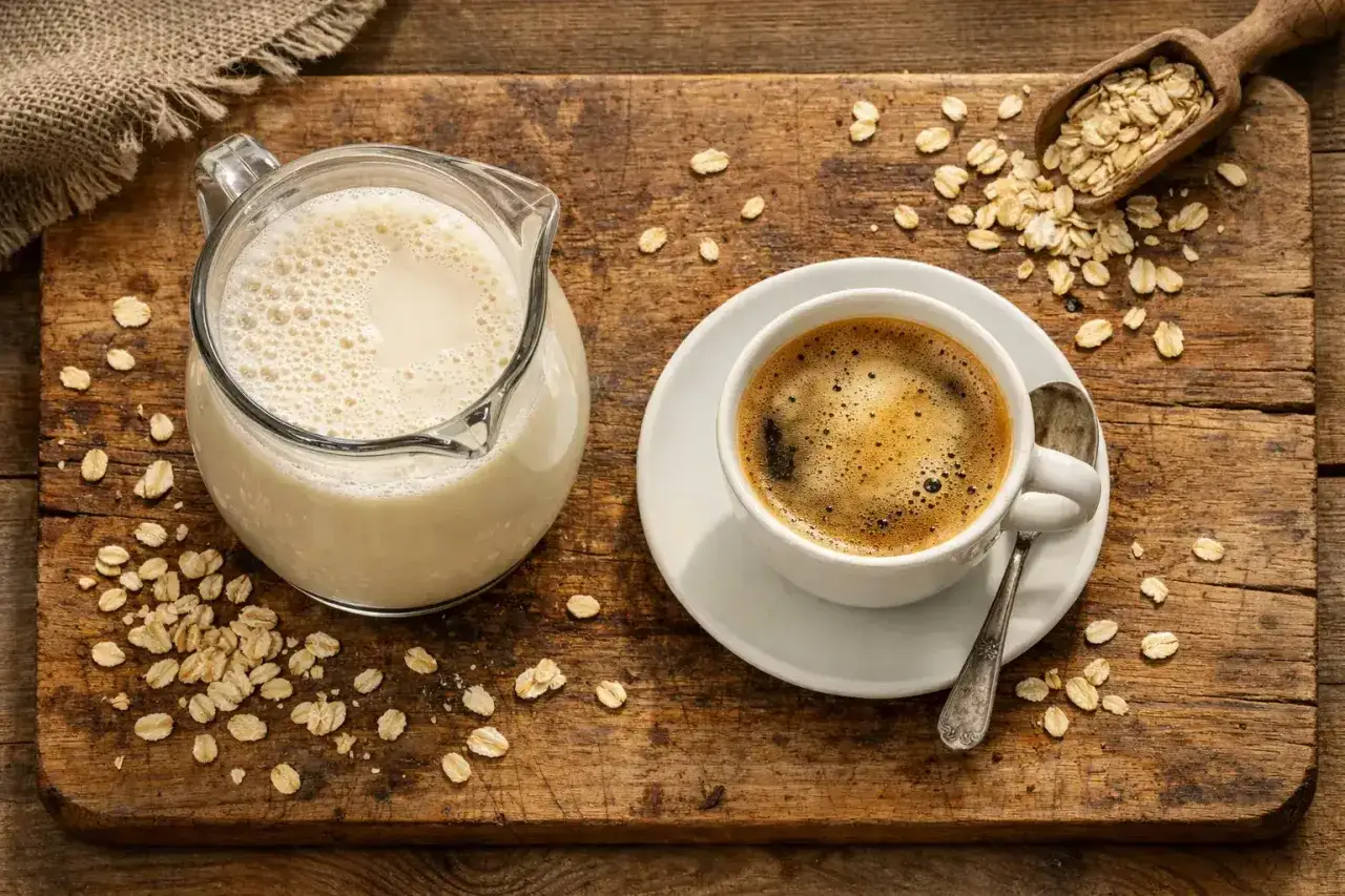 Eine Kanne Hafermilch und eine Tasse Kaffee stehen auf einem Holzbrett. Haferflocken sind verstreut. Kaffee mit Hafermilch Kalorien sind oft geringer.