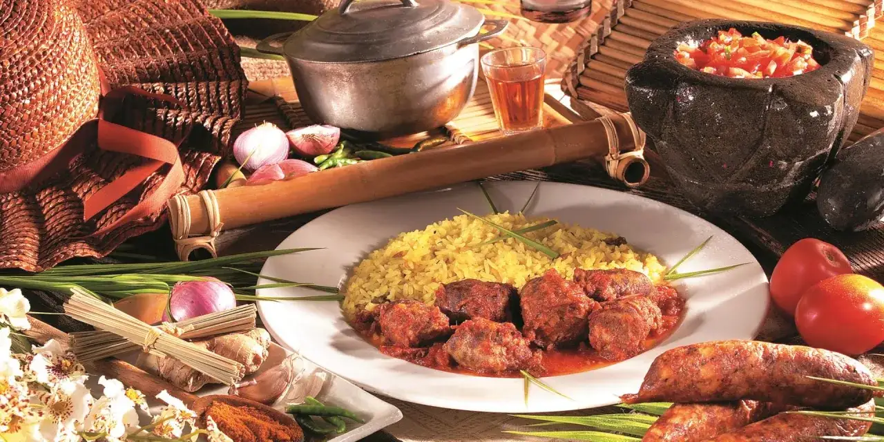 Un plat de rougail saucisses traditionnel, servi avec du riz jaune. Des ingrédients frais comme des oignons, du gingembre et des tomates sont disposés autour.