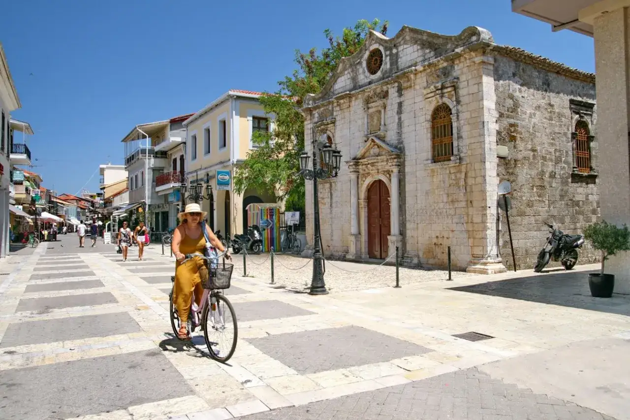 Kobieta na rowerze przemierza ulicę w Lefkada miasto, obok zabytkowej kamiennej budowli.
