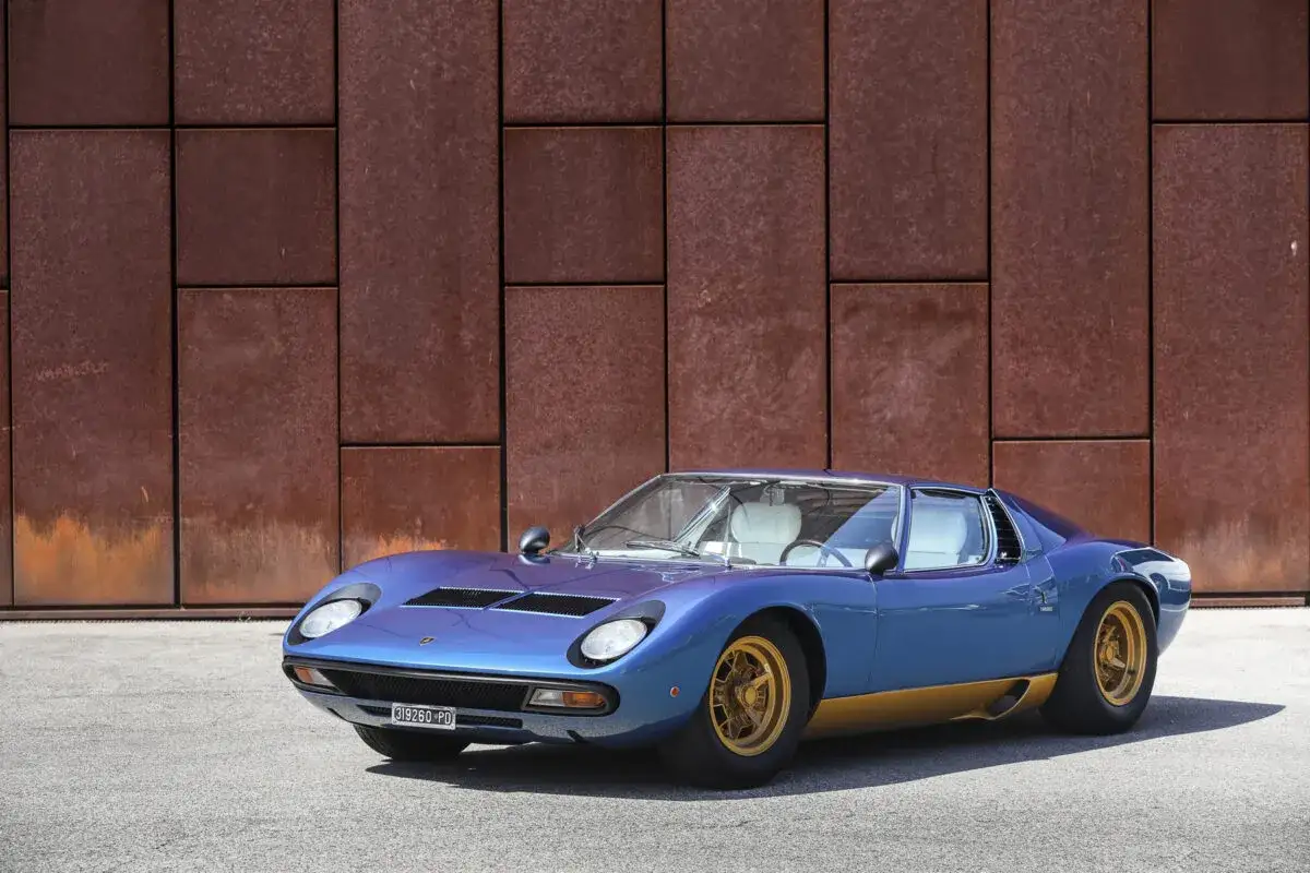 Ein blauer Lamborghini Miura S mit goldenen Felgen steht vor einer rostigen Metallwand.