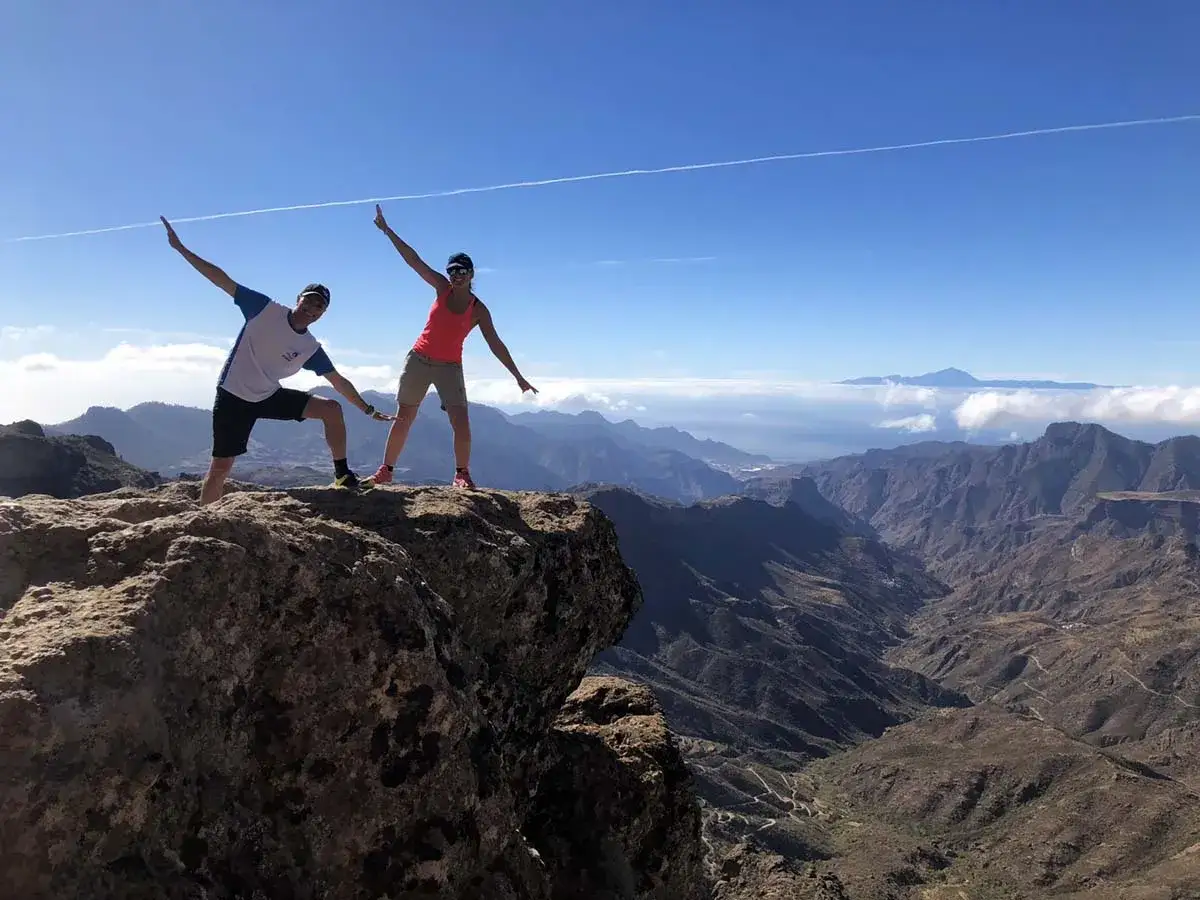 Dos excursionistas celebran en la cima de una montaña en Gran Canaria, con un paisaje espectacular de barrancos y el mar al fondo.