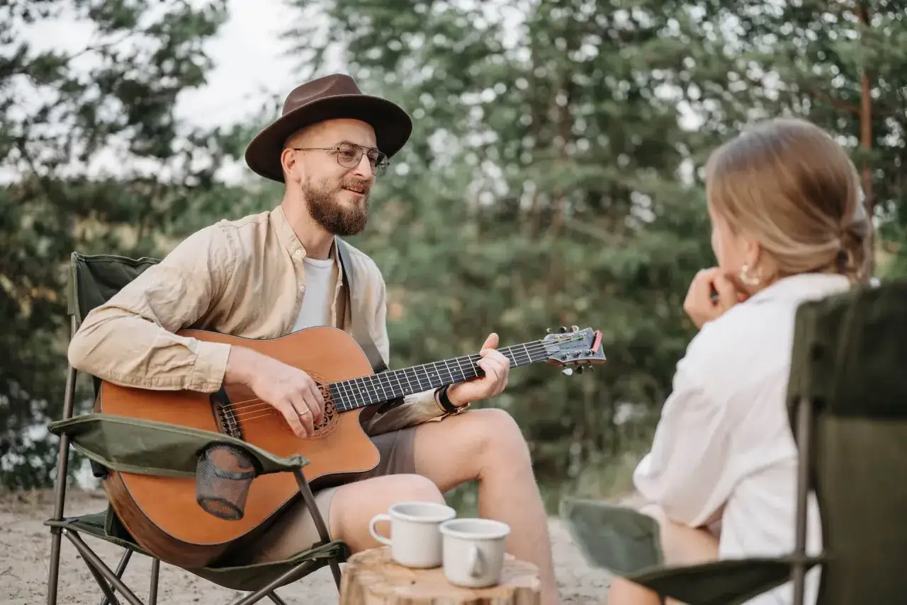 Mann mit Hut spielt Akustikgitarre für Frau im Freien. Vielleicht lernt er gerade, wie man Akustikgitarre lernt und teilt seine Fortschritte.